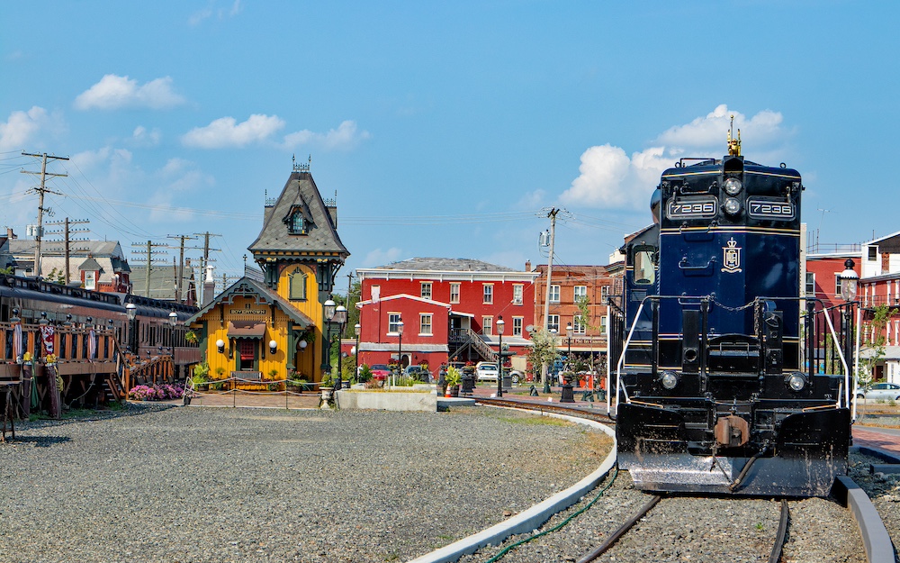 A dark blue diesel locomotive next to a yellow depot with brick buildings beyond