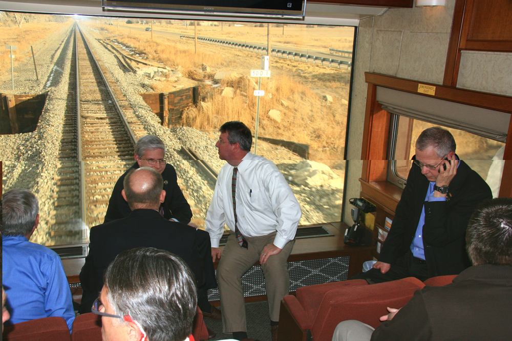 People talking in railroad car with large glass window looking at track in background