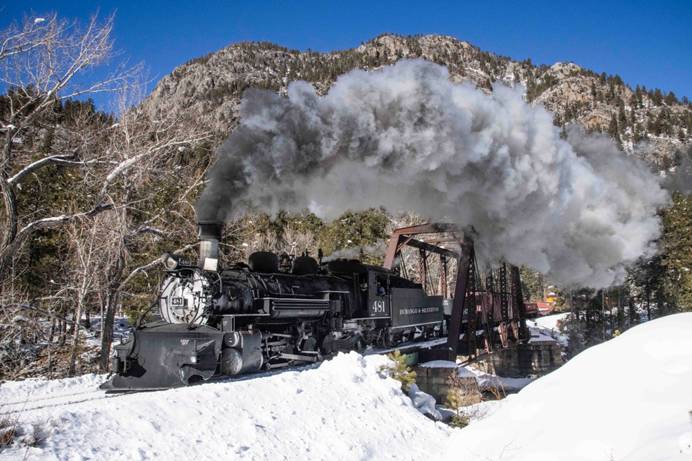 Steam engine with train crossing bridge in snowy landscape.