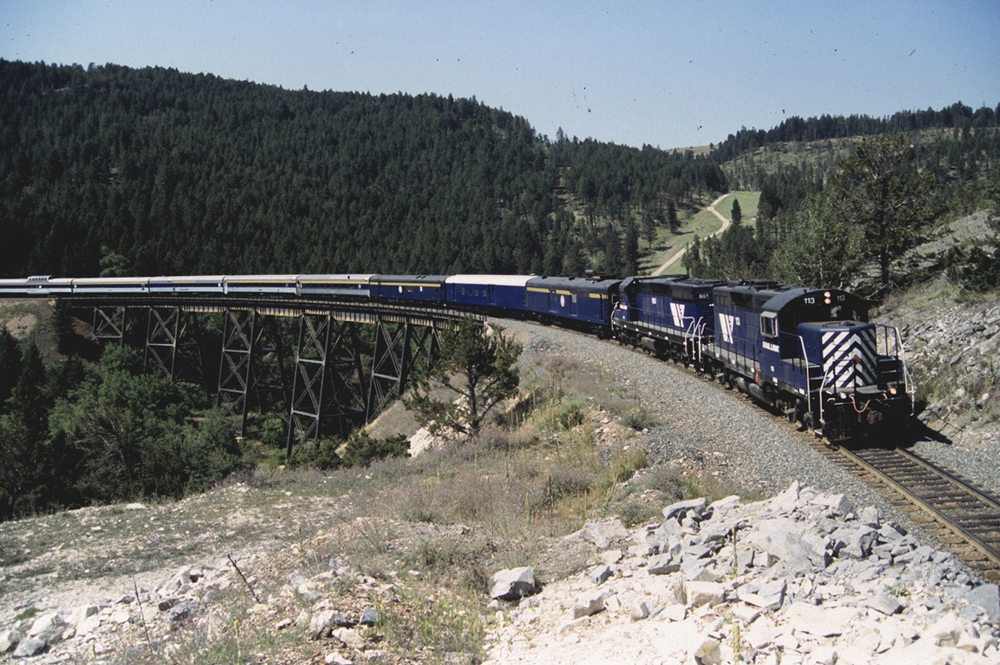 Passenger train with blue freight locomotives crossing bridge in mountains