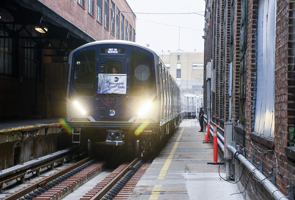 Subway train with "inaugural run" banner moves between buildings at yard complex