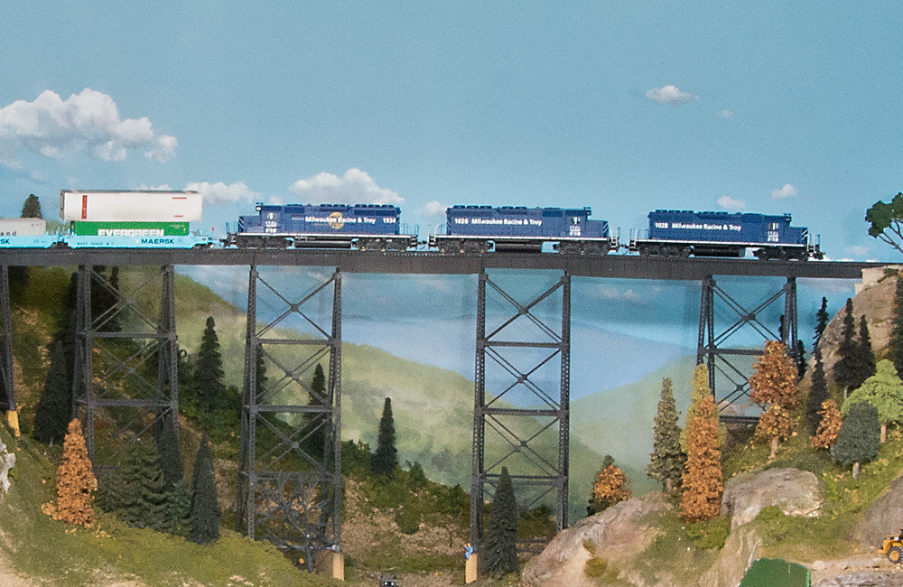 Three blue HO scale diesels pull a stack train over a high steel trestle