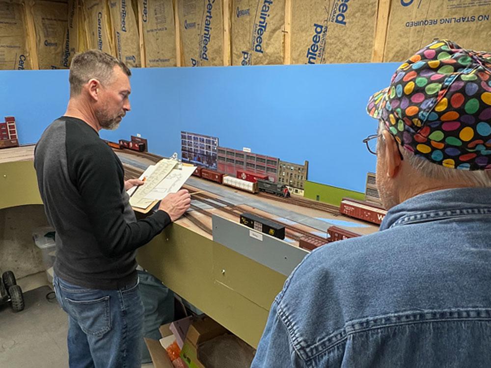 A man in a polka-dot cap at right watches another man operate a model railroad
