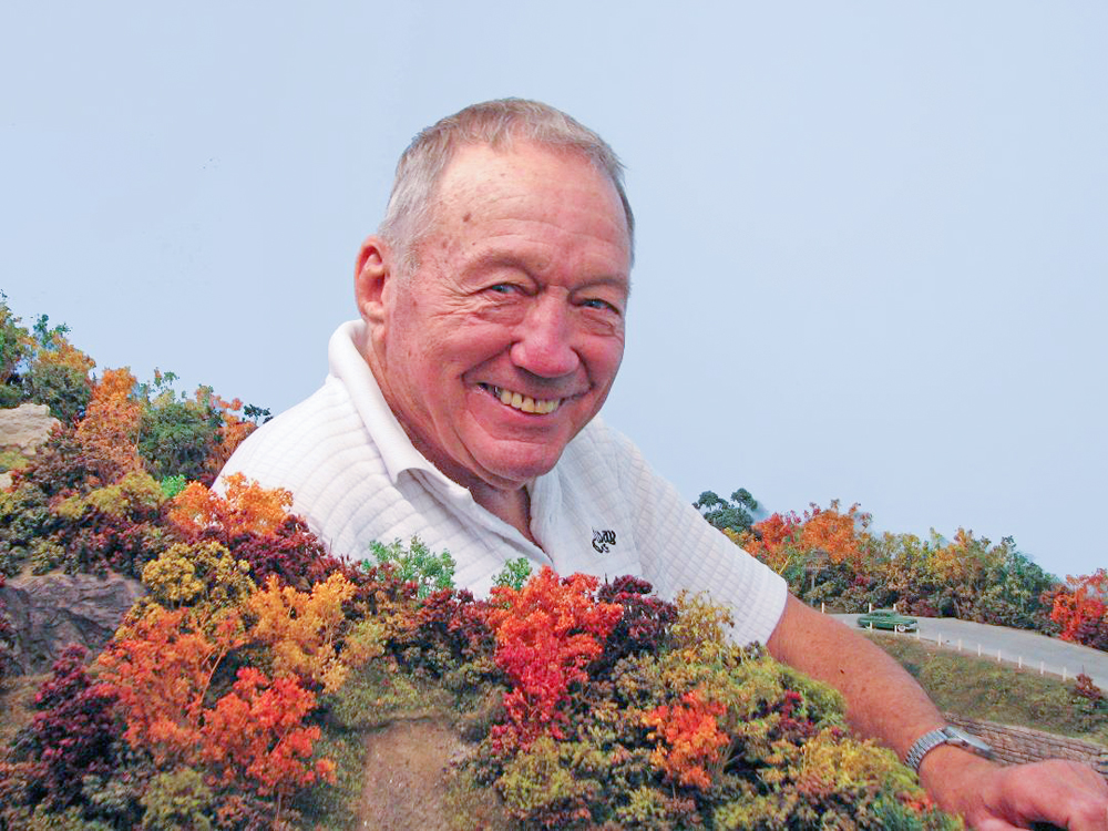 Color photo of smiling man standing by model railroad layout.