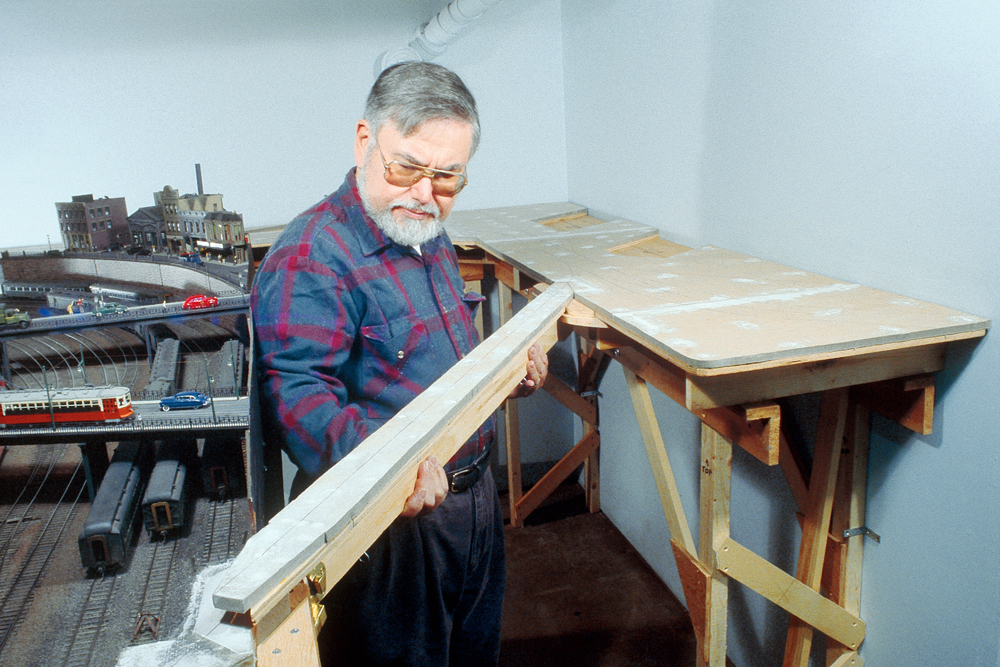 Color photo of man with gray hair and beard wearing glasses holding benchwork.