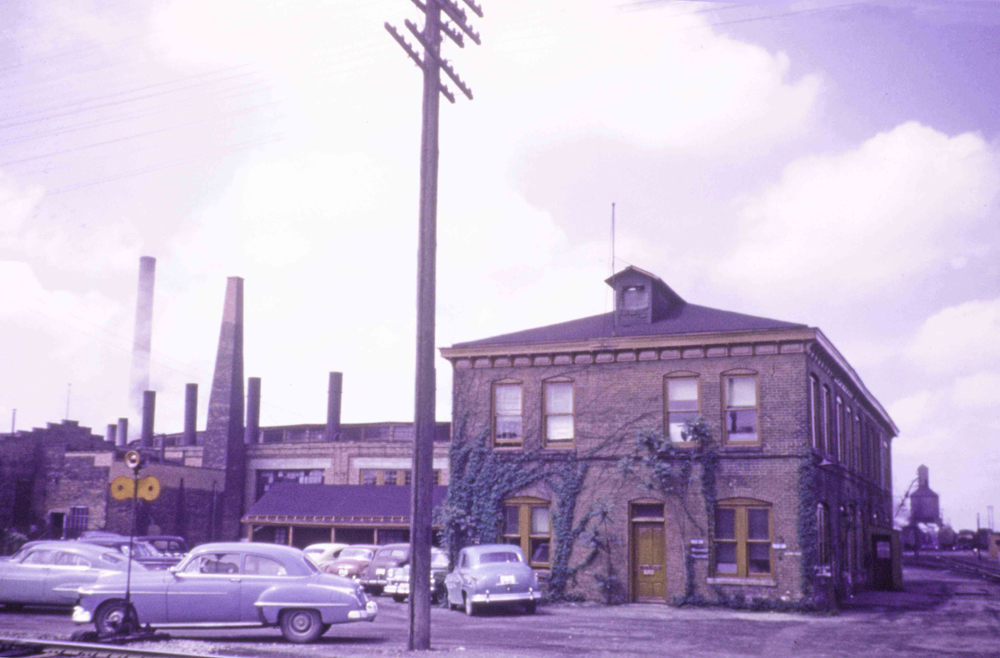 Color photo of two-story brick railroad building with vehicles in foreground.