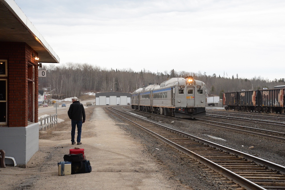 Under overcast skies a silver and blue two car passenger train reverses past a red brick depot