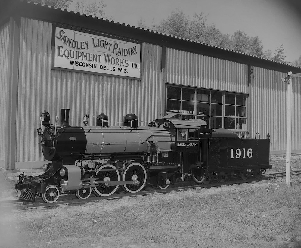 Black-and-white photo of grand-scale, 4-4-2 steam locomotive in front of a shop building.