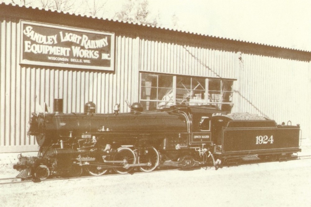 Black-and-white photo of grand-scale 4-6-2 steam locomotive in front of a shop building.