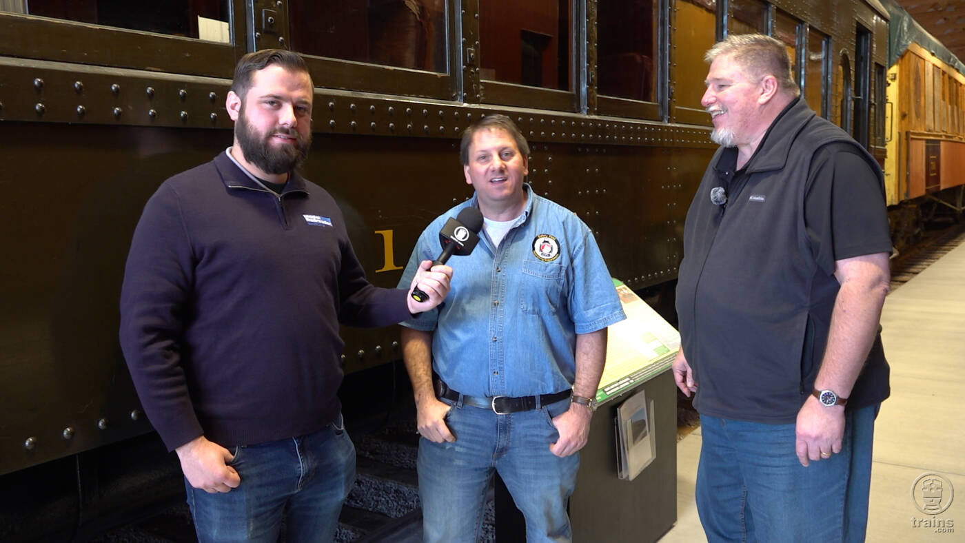 Three men standing next to a motorcar for a TV interview. Trains LIVE — Restoring Montana Western No. 31