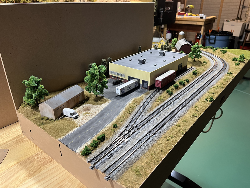 Small model train set up with gray and black track, green grass and trees, dark gray roads, miniature vehicles, and two buildings of different sizes.