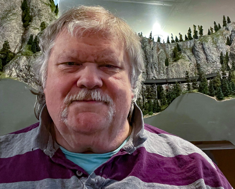 Color photo of man with moustache standing in front of model railroad layout.