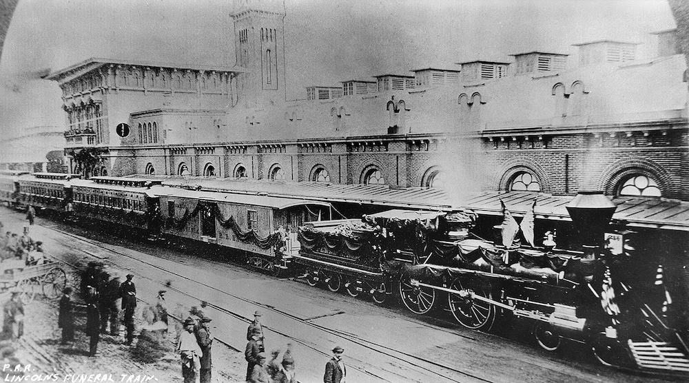 Black and white photo of steam-powered passenger train, draped in funeral attire, at the station.