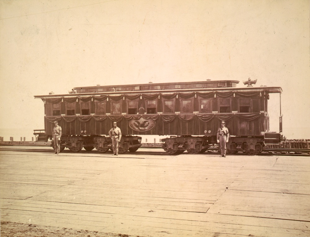 Black and white photo of passenger car draped in funeral attire being guarded.