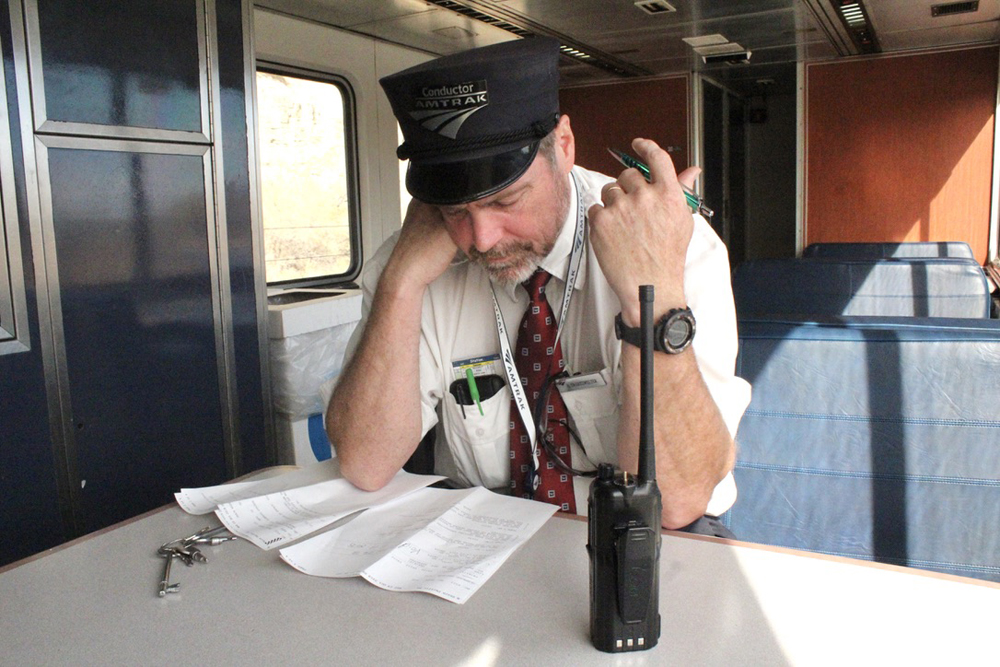 Amtrak conductor with head down over paperwork at table on board train