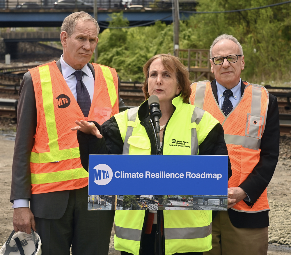Woman speaks at podium with two men standing behind her. All three are in hi-visibility vests