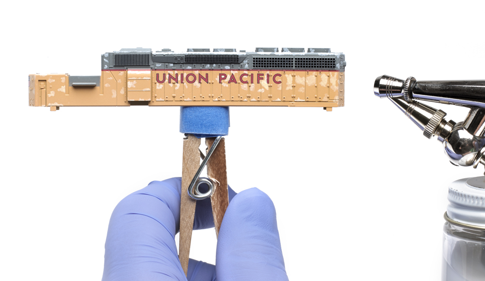 Color photo of gloved hand holding a clothespin and locomotive shell with an airbrush visible at right edge of the image.