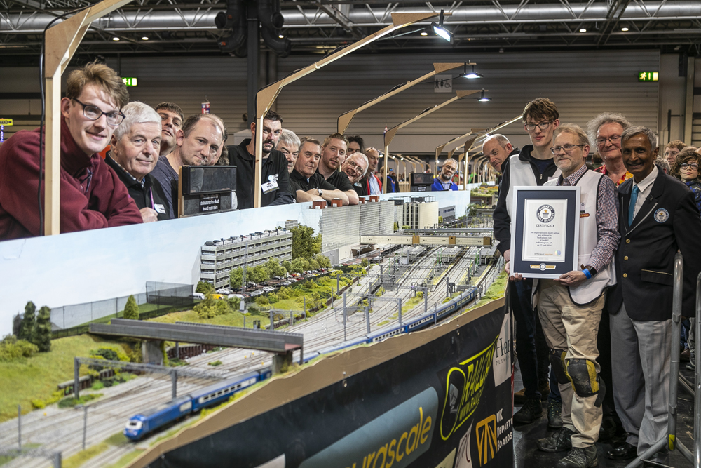 A goup of people stand around a part of a model railroad layout, with one man holding a world record certificate