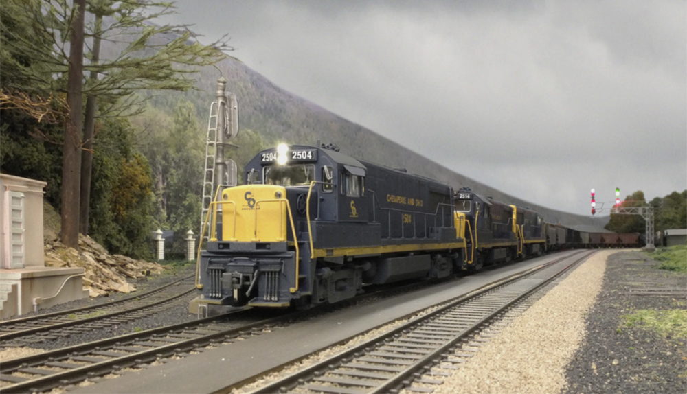 A model locomotive in a blue and yellow paint scheme in front of a mountainous backdrop