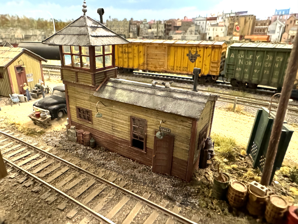 Small, model railroaded structure including second floor tower with lighted windows, gray pointed roof, and brown and yellow boards and trim.