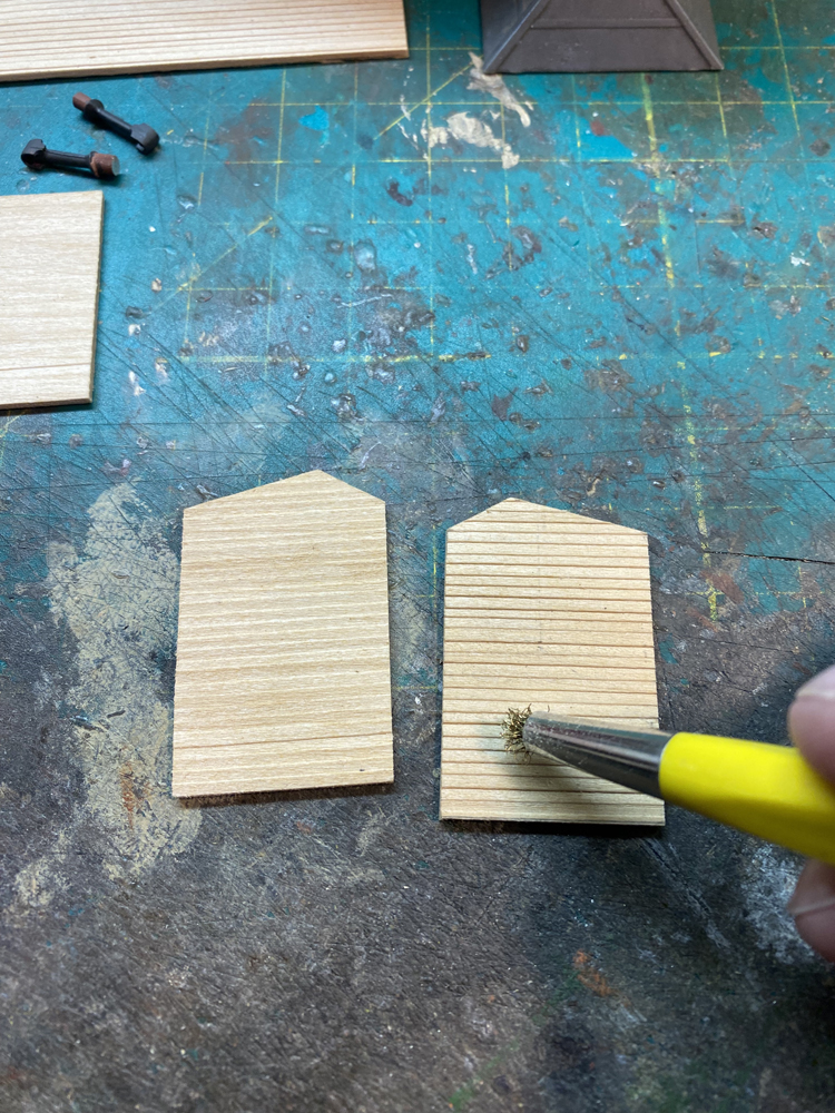 Small pieces of wood for modeling lay flat on worn green work mat with yellow and silver brush with brass-colored bristles.