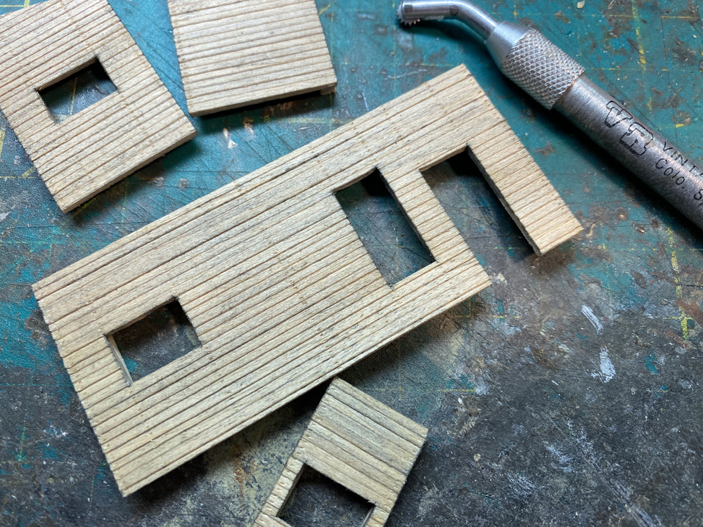 Brown rectangular pieces of wood representing model structure walls with siding, window, door, and nail details.