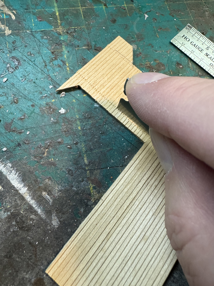A tan hand holding a single-edge razor blade on a brown piece of model wood siding along a line of simulated nail holes.