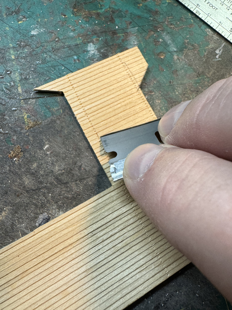 A tan hand holding a silver single-edge razor blade on a brown piece of model wood siding parallel to one of the siding boards.