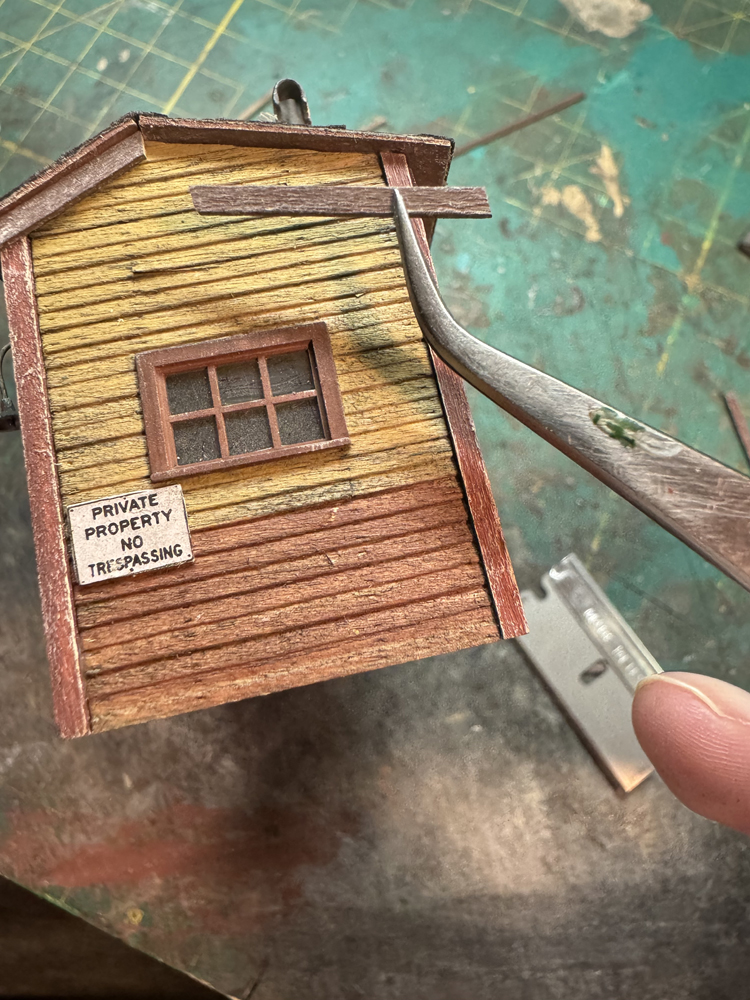Model railroad building with yellow and red faded paint, as well as silver tweezer holding small red trim board.