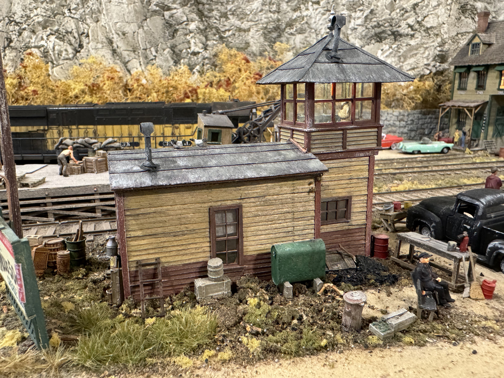 Model railroad building with yellow and red faded paint, as well as silver tweezer holding small red trim board.