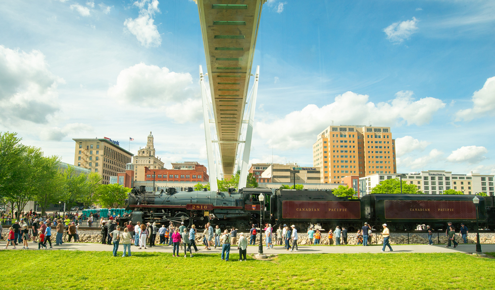 Steam engine surrounded by people parked under pedestrian bridge