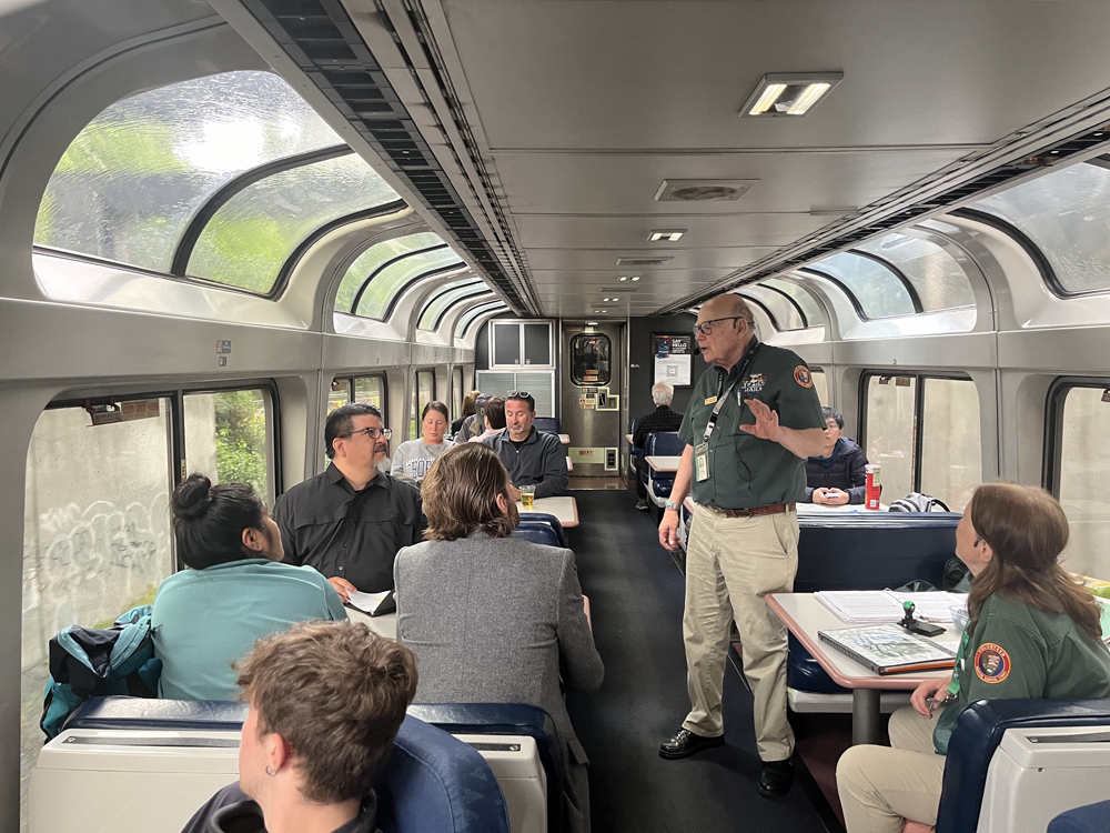 Man standing in lounge car, talking to others at table