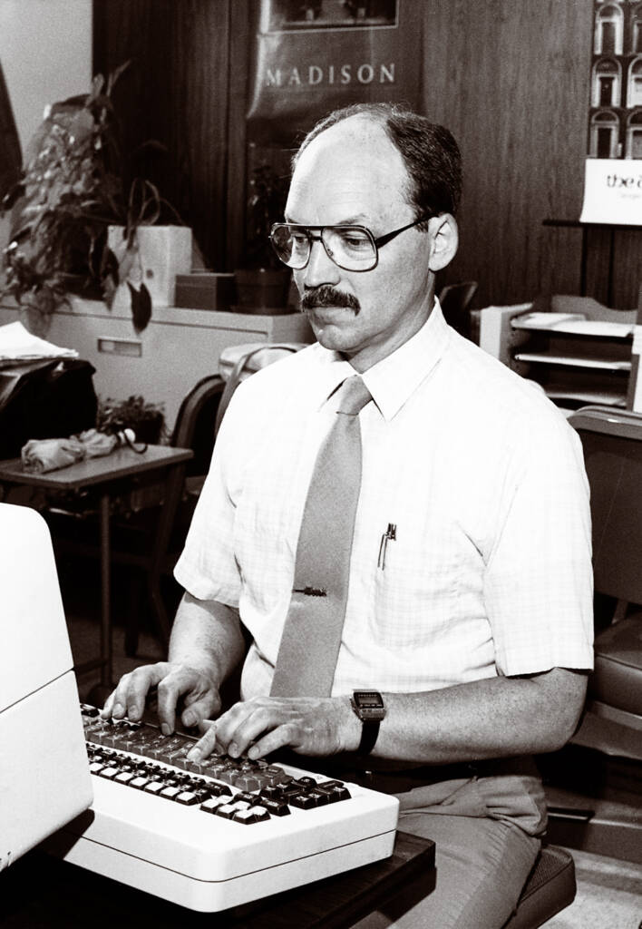 Black-and-white photo of man wearing glasses, dress shirt, and necktie typing at a word processor.