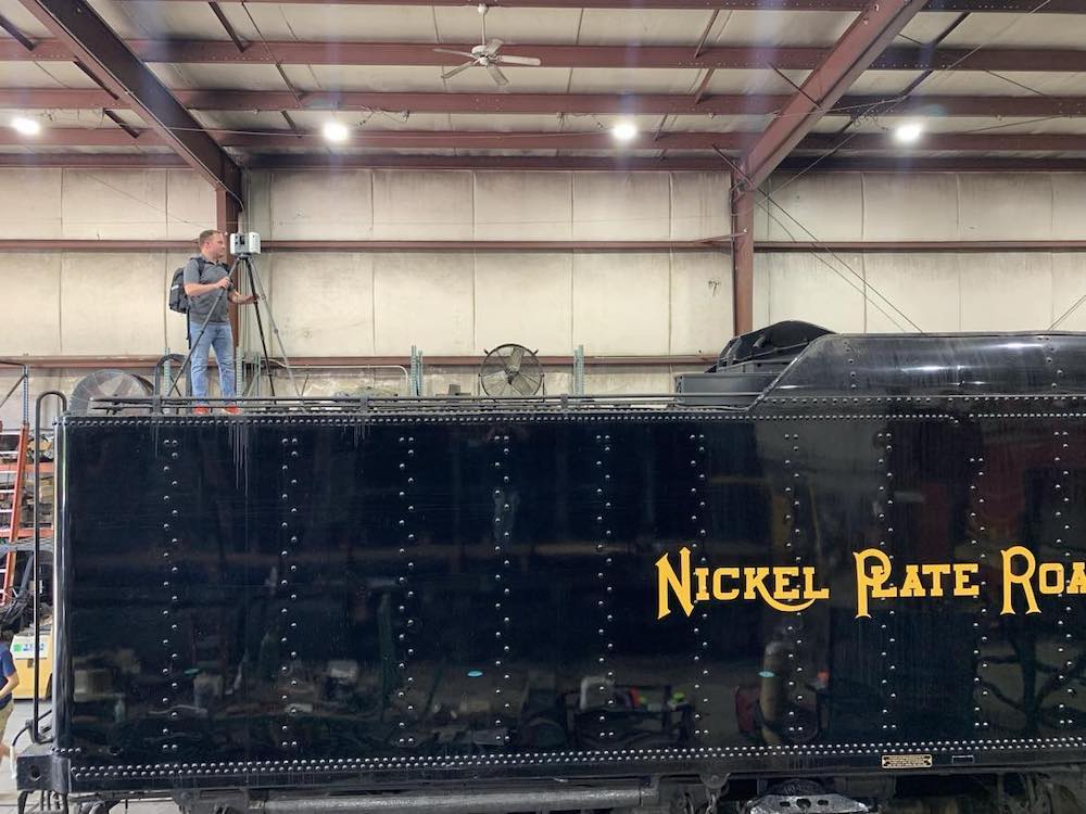 Man with tripod and scanning machine stands on top of steam locomotive tender.