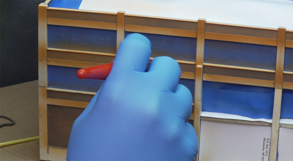 A blue-gloved hand airbrushes brick red paint on a plastic model of a factory