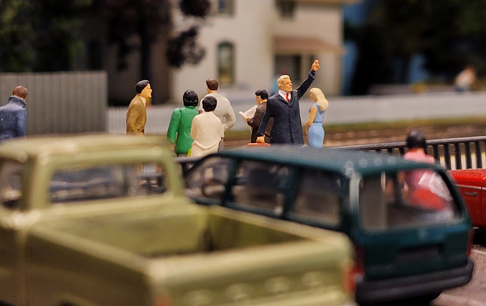 A model figure in a blue coat raises his arm on a model railroad layout
