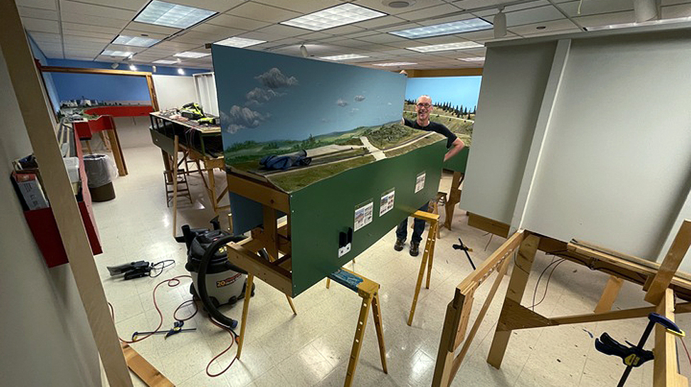 A model railroad layout being disassembled with a man standing between sections of the deconstructed layout