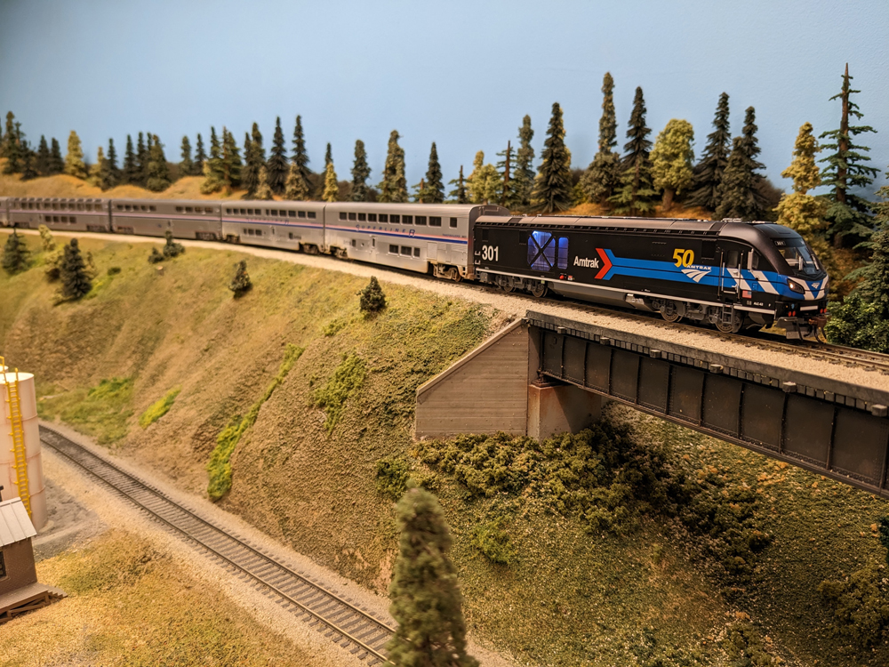 A black model locomotive leads a silver model passenger train across a bridge on a model railroad layout