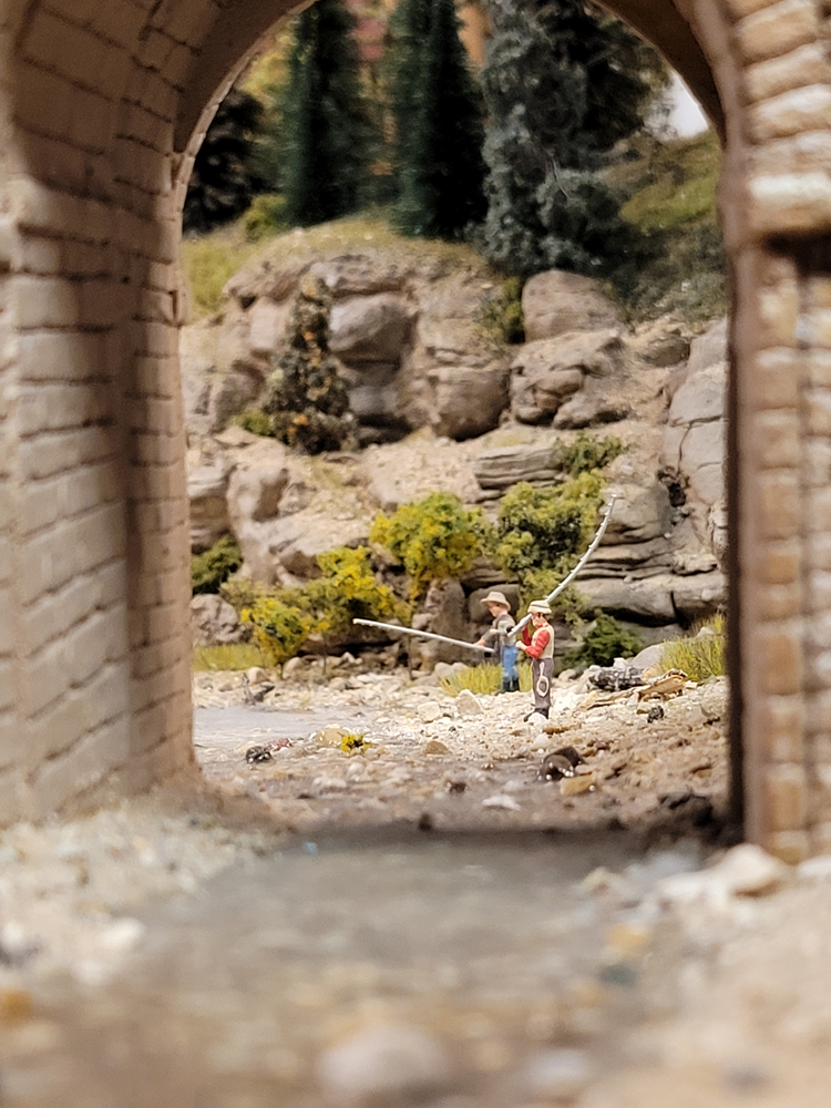 Two model figures stand with fishing poles under a bridge on a model railroad layout