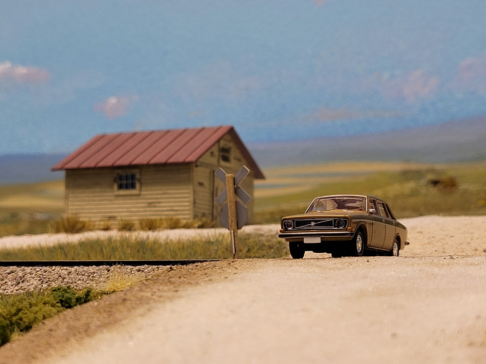 A gold model sedan next to a set of model train tracks on a model railroad layout