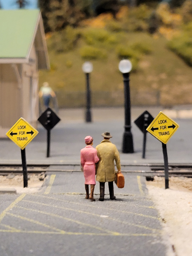 Two figures stand at a railroad crossing on a model railroad layout