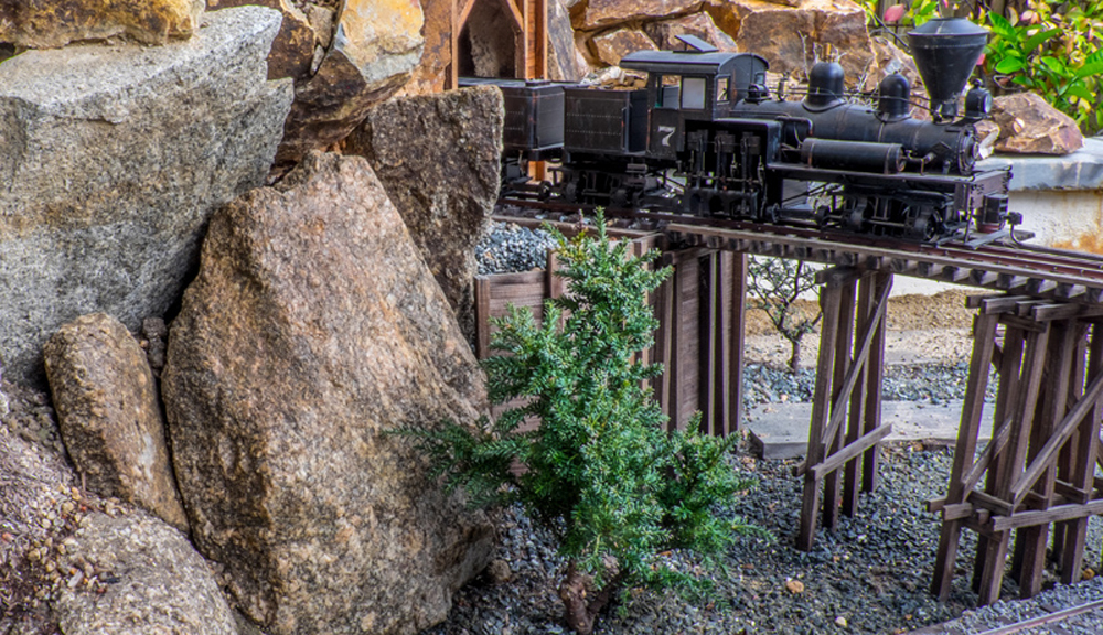 A toy train in an outdoor setting on an elevated trestle bridge with a rock formation at the left of the image
