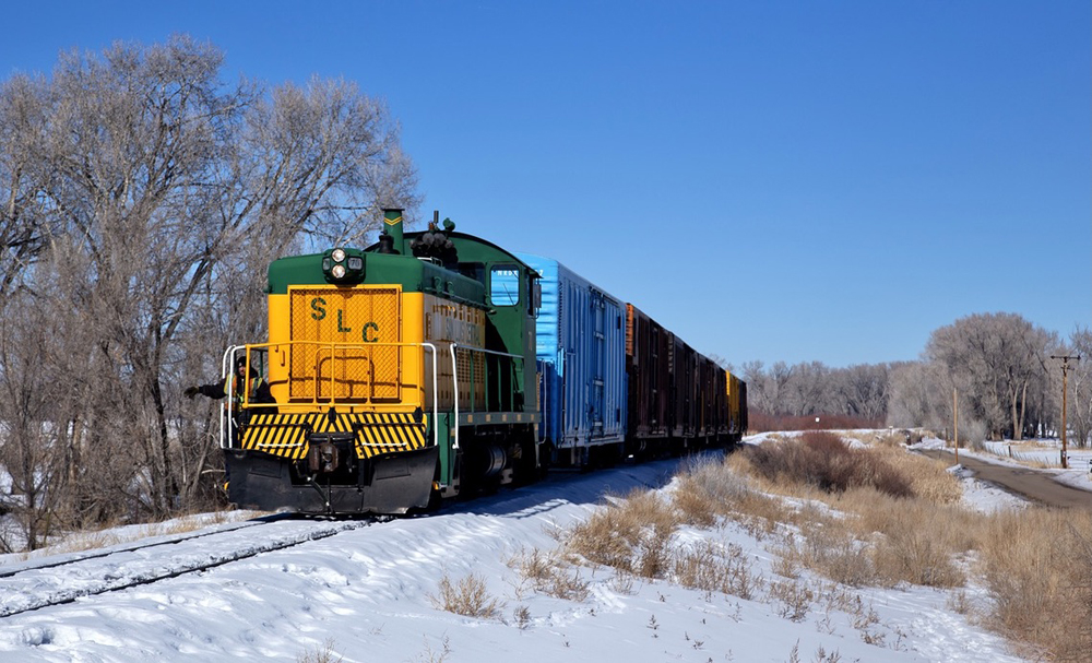 Green and yellow locomotive with short string of cars on straight track in snow.