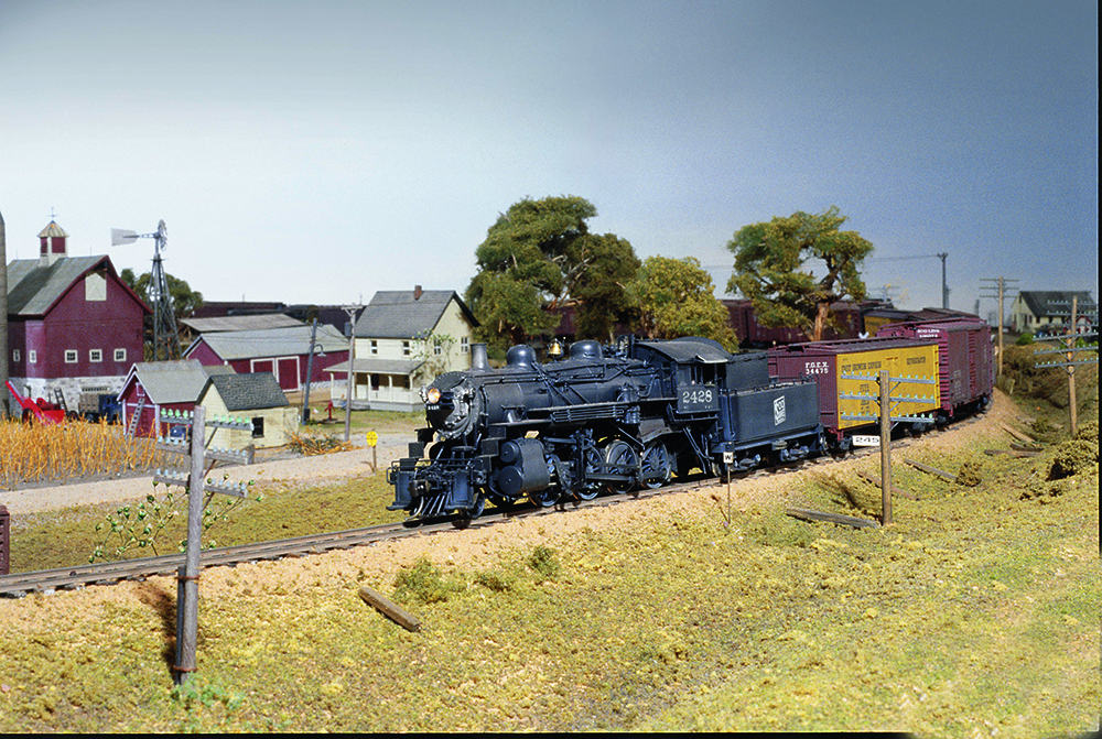 Black model steam locomotive pulling a short train of colorful cars past a rural farm scene during a summer afternoon.