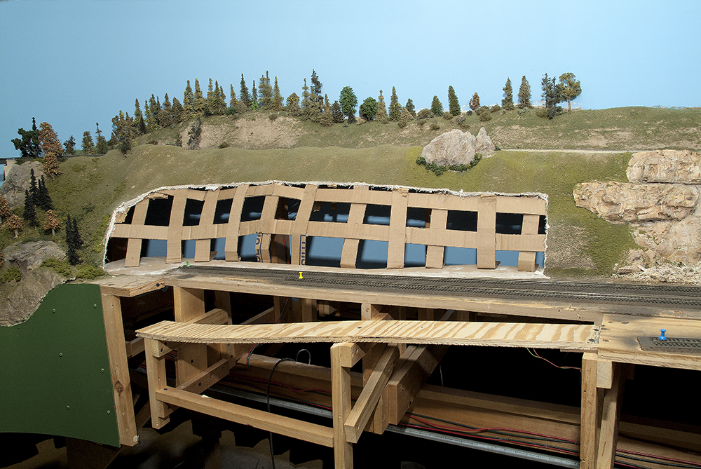 A green hillside on a model train set with part cut away to show the supporting lattice of brown cardboard strips.
