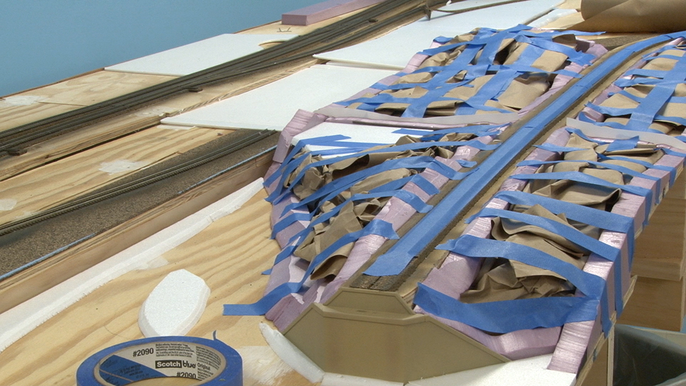 Model train hill supports on a toy train layout, made with strips of pink foam and blue masking tape and stuffed with crumpled brown paper.