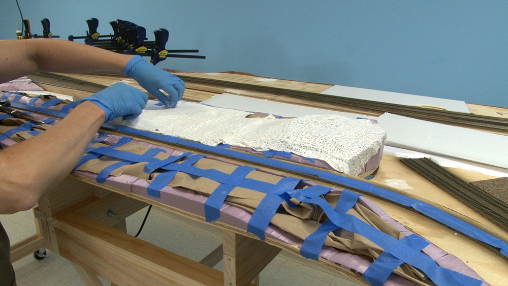 Hands wearing blue plastic gloves placing white plaster sheets over model train scenery made from pink insulation board, brown crumpled paper, and blue masking tape.
