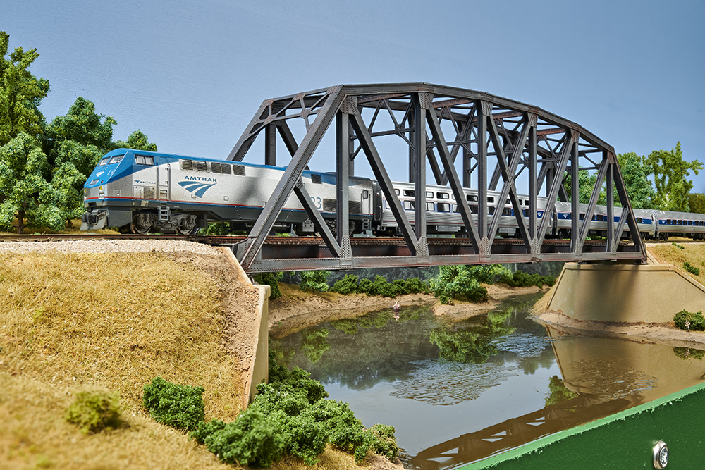Blue and silver model train crossing a brown steel bridge over a glossy, rural river lined with green trees, tall grass, and bushes.