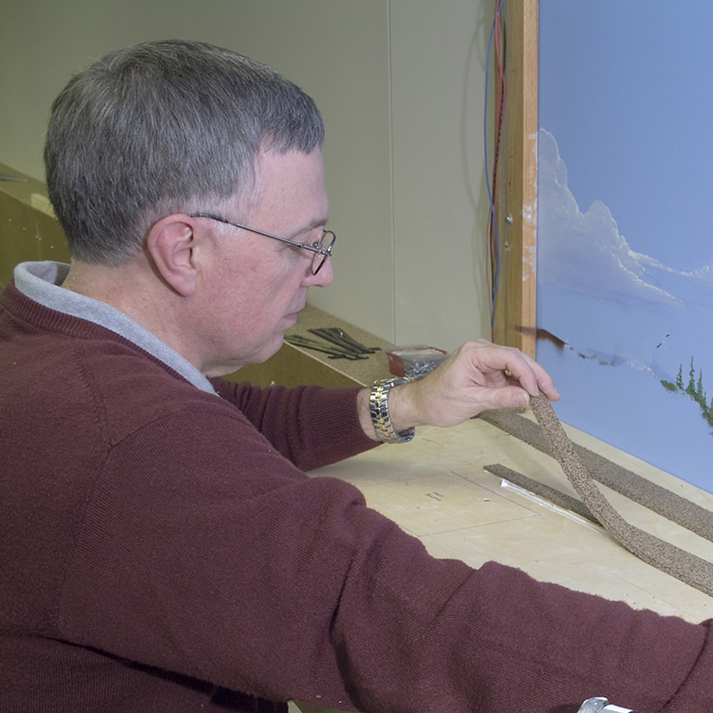 Color photo of man in dark red sweater attaching cork strip to model railroad.