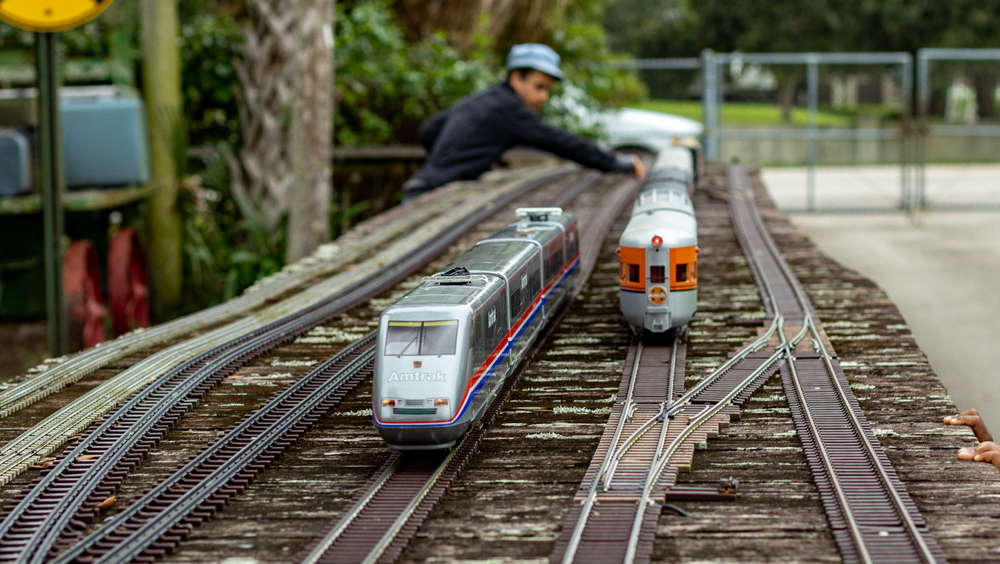 An individual is seen in the background of the image manipulating the train layout while two model trains approach the camera on the layout